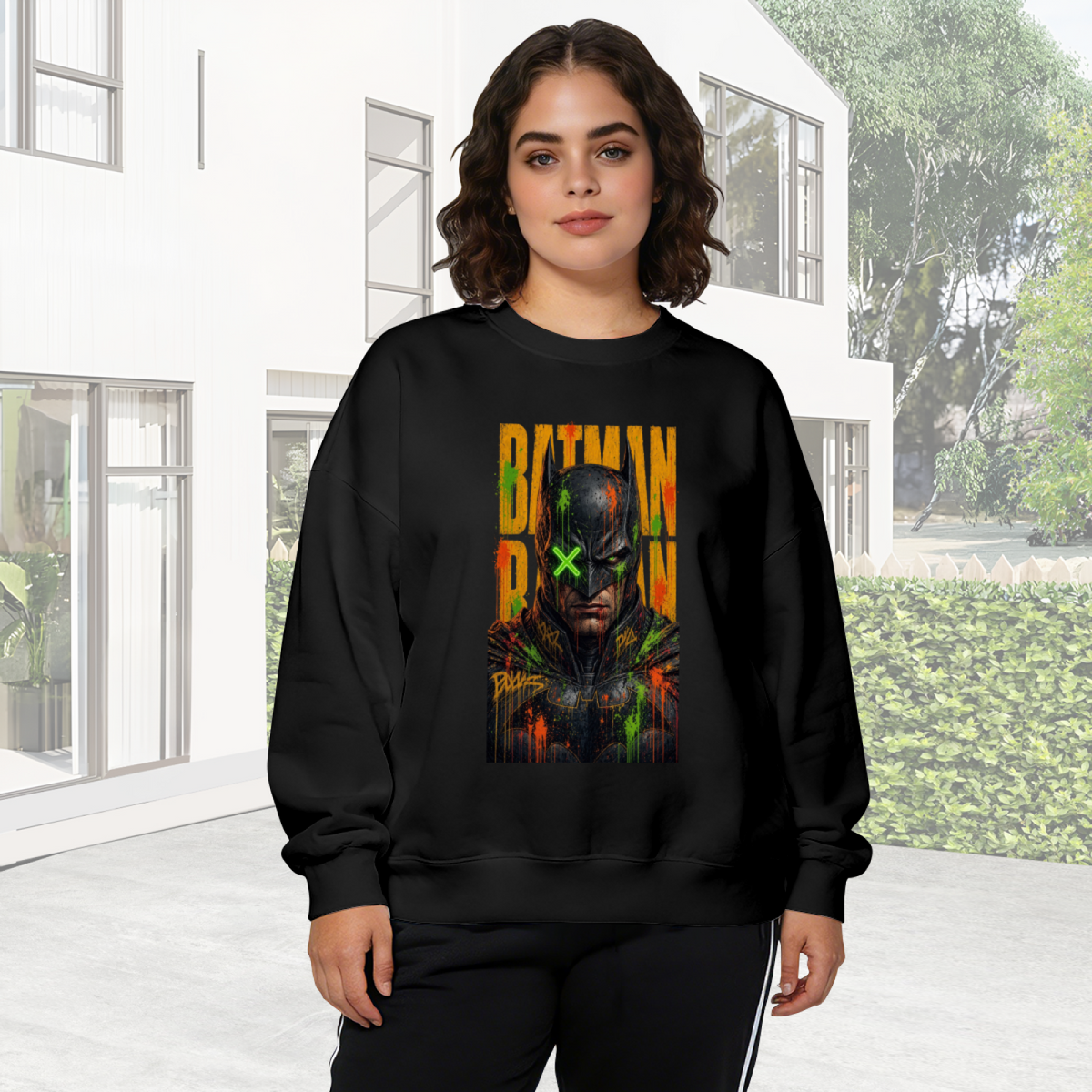 Outdoor photograph of a model in a women's crewneck sweatshirt bearing a tall graffiti-style cowl portrait with a green X across the eyes